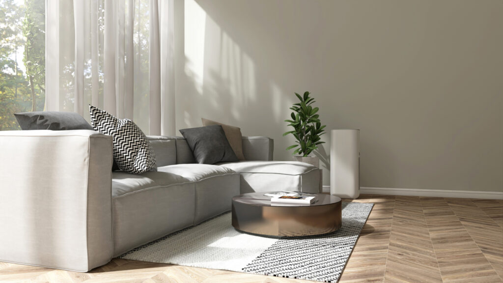 A modern living room with a light gray sofa adorned with patterned cushions. A round black coffee table sits on a textured rug over herringbone wooden flooring. A few green plants decorate the space near large windows that let in ample sunlight.